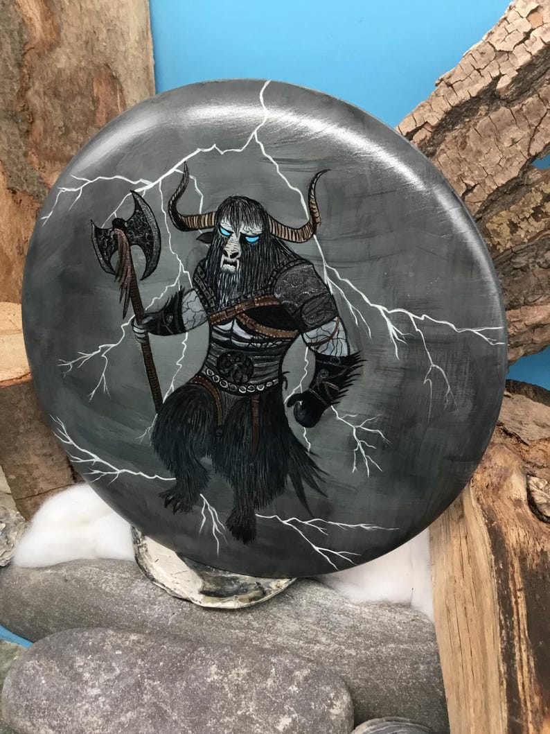 May include: A circular, grey painted wooden plaque depicting a Minotaur warrior with an axe, set against a backdrop of lightning. The Minotaur has blue eyes and black fur. The plaque is displayed on a stone surface.