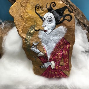 Op de afbeelding: Handgeschilderde rotskunst met een figuur in een rood-gouden jasje, witte gefranjerde kraag en een zwart-witte hoed. De figuur houdt een klein kopje en een lange, dunne pijp vast. De rots heeft een goud-bruine achtergrond.