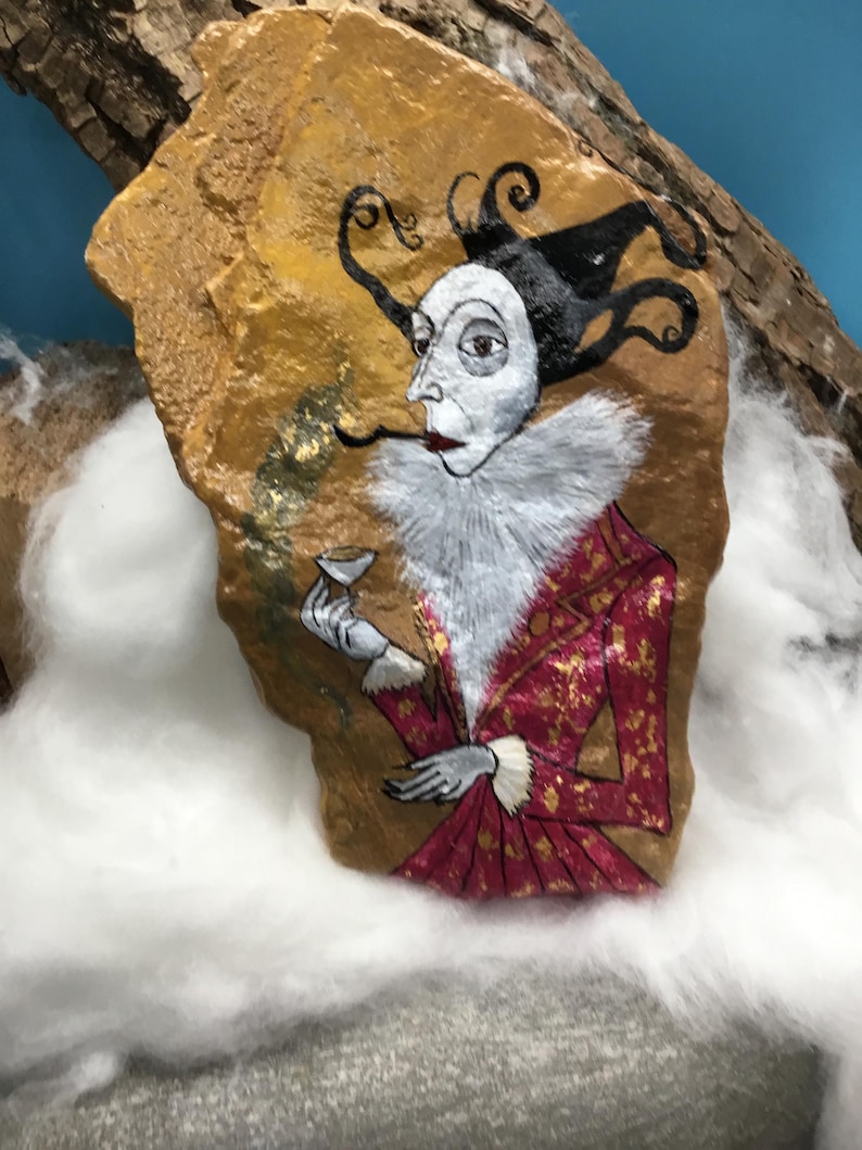 Op de afbeelding: Handgeschilderde rotskunst met een figuur in een rood-gouden jasje, witte gefranjerde kraag en een zwart-witte hoed. De figuur houdt een klein kopje en een lange, dunne pijp vast. De rots heeft een goud-bruine achtergrond.