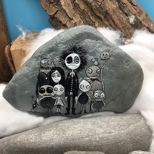 Puede incluir: Una piedra gris lisa con una ilustraci&oacute;n detallada pintada a mano de un grupo de personajes estilizados con ojos grandes y rasgos oscuros. La obra de arte muestra una figura central alta rodeada por varias figuras m&aacute;s peque&ntilde;as.