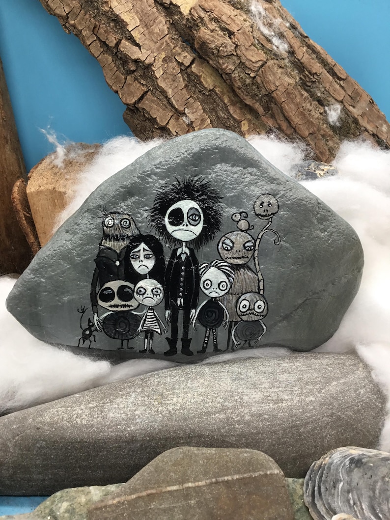 Puede incluir: Una piedra gris lisa con una ilustraci&oacute;n detallada pintada a mano de un grupo de personajes estilizados con ojos grandes y rasgos oscuros. La obra de arte muestra una figura central alta rodeada por varias figuras m&aacute;s peque&ntilde;as.