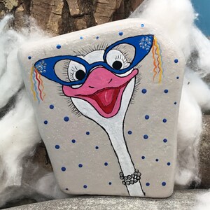 May include: A hand-painted rock featuring an ostrich wearing blue glasses with rhinestones and a pearl necklace. The background is a light grey with blue polka dots.