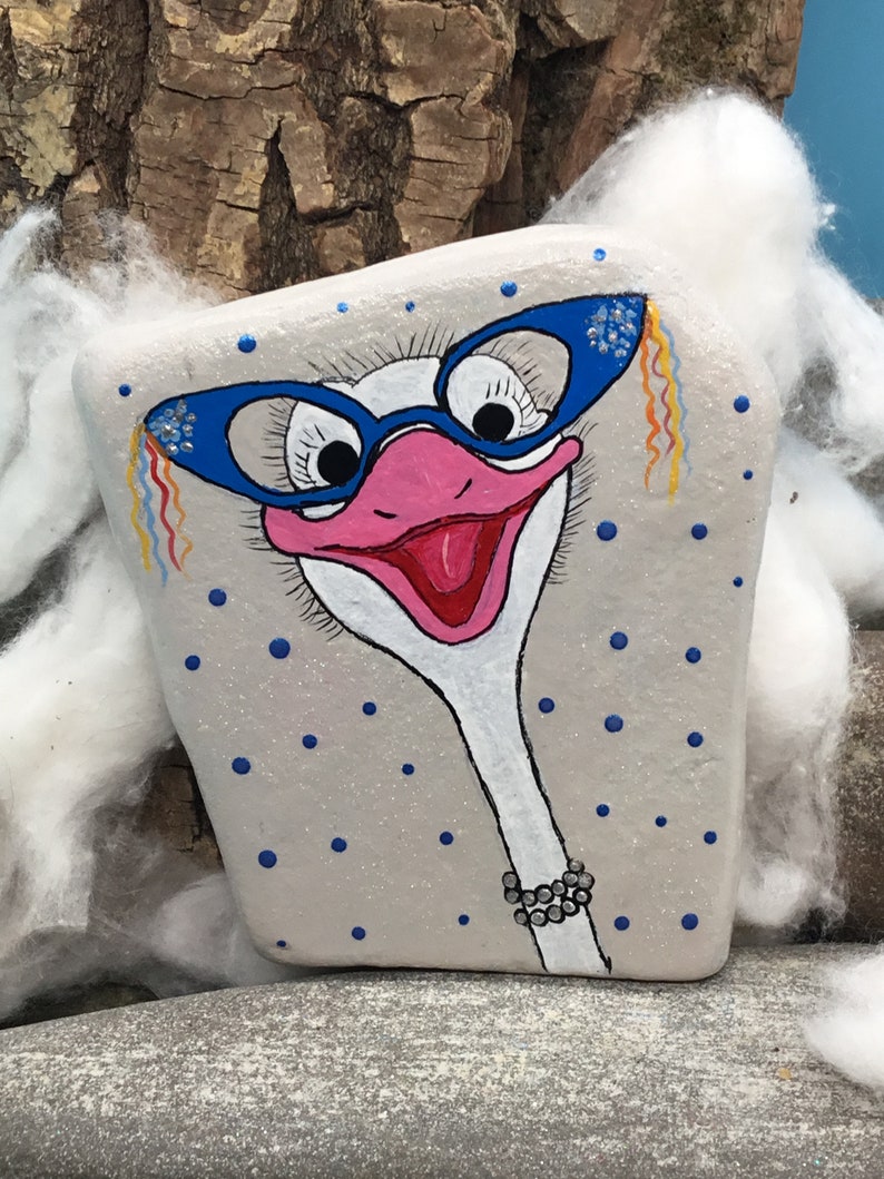 May include: A hand-painted rock featuring an ostrich wearing blue glasses with rhinestones and a pearl necklace. The background is a light grey with blue polka dots.