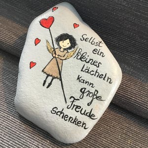 K&ouml;nnte beinhalten: Handbemalter Stein mit einer Illustration eines Engels, der einen herzf&ouml;rmigen Ballon h&auml;lt. Der Stein ist hellblau mit roten Herzakzenten und schwarzer Schrift, die lautet: &bdquo;Schon ein kleines L&auml;cheln kann gro&szlig;e Freude schenken&ldquo;.