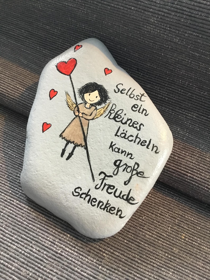 K&ouml;nnte beinhalten: Handbemalter Stein mit einer Illustration eines Engels, der einen herzf&ouml;rmigen Ballon h&auml;lt. Der Stein ist hellblau mit roten Herzakzenten und schwarzer Schrift, die lautet: &bdquo;Schon ein kleines L&auml;cheln kann gro&szlig;e Freude schenken&ldquo;.
