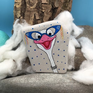 May include: Hand-painted rock art of an ostrich wearing blue glasses and a pearl necklace. The ostrich is painted on a grey rock with white polka dots. The rock is sitting on a bed of white cotton.