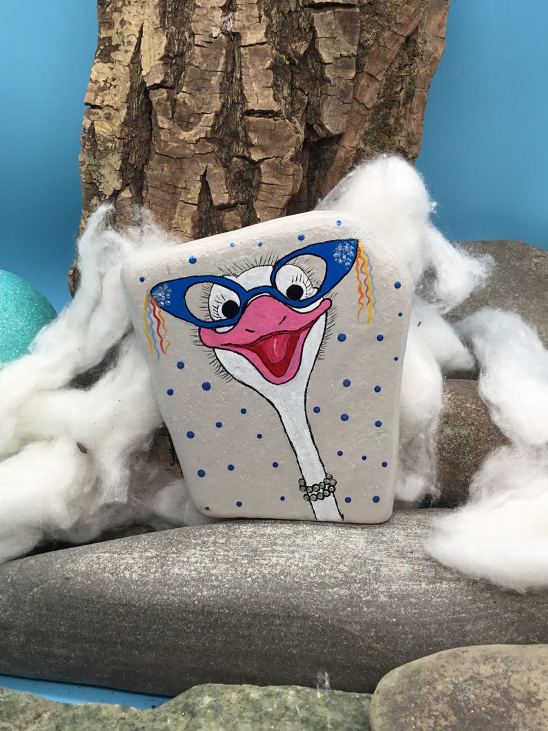 May include: Hand-painted rock art of an ostrich wearing blue glasses and a pearl necklace. The ostrich is painted on a grey rock with white polka dots. The rock is sitting on a bed of white cotton.