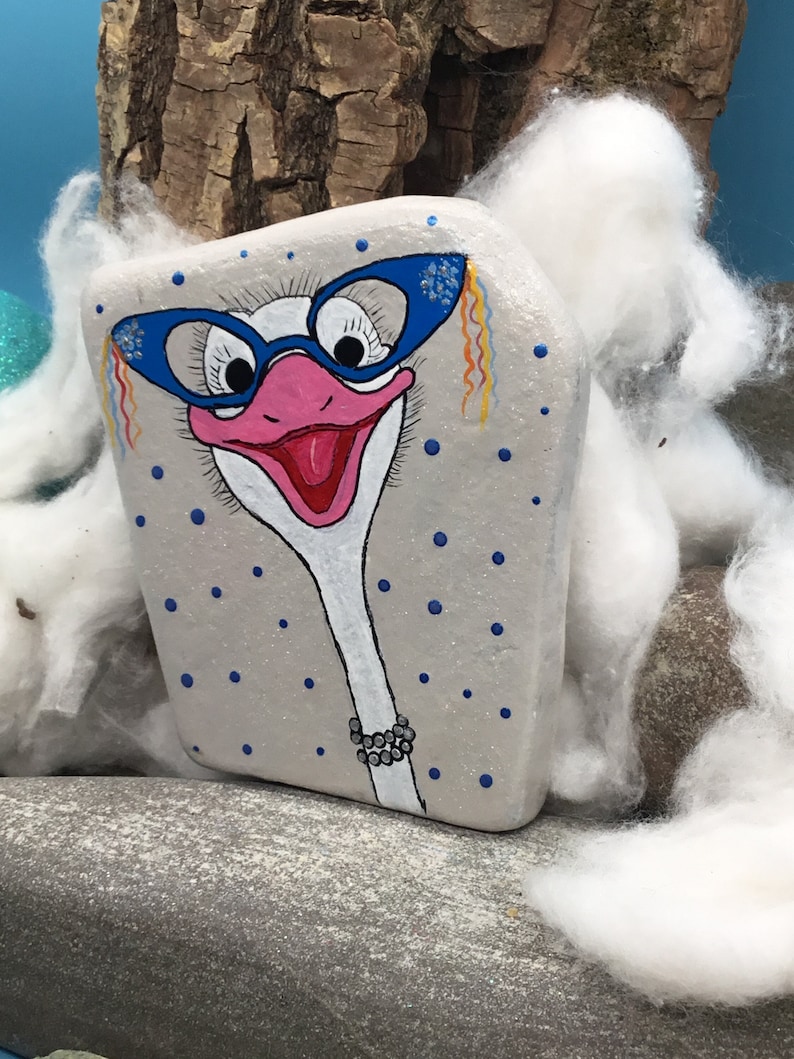 May include: Hand-painted rock art of an ostrich with blue glasses, a pink beak, and a white neck with a pearl necklace. The rock is painted with a light grey background and blue polka dots.