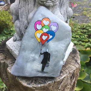 May include: A hand-painted, gray stone features a whimsical illustration of a person holding colorful heart-shaped balloons. The balloons are in shades of yellow, orange, red, green, purple, and blue. The person is dressed in a black coat and carrying a briefcase.