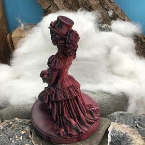 May include: A 3D printed figurine of a person wearing a top hat and a long, layered dress with a ruffled skirt. The figure is colored in a deep red and black gradient, standing on a circular base. The design features intricate textures.