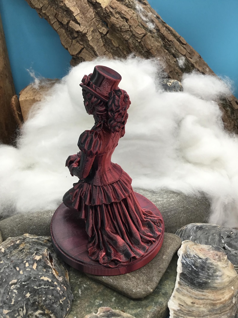 May include: A 3D printed figurine of a person wearing a top hat and a long, layered dress with a ruffled skirt. The figure is colored in a deep red and black gradient, standing on a circular base. The design features intricate textures.