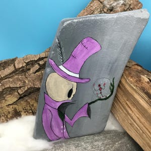 May include: Hand-painted rock art featuring a whimsical skeleton figure in a purple top hat and coat, holding a crystal ball. The artwork is set against a gray background with a blue sky and wood accents, creating a unique decorative piece.