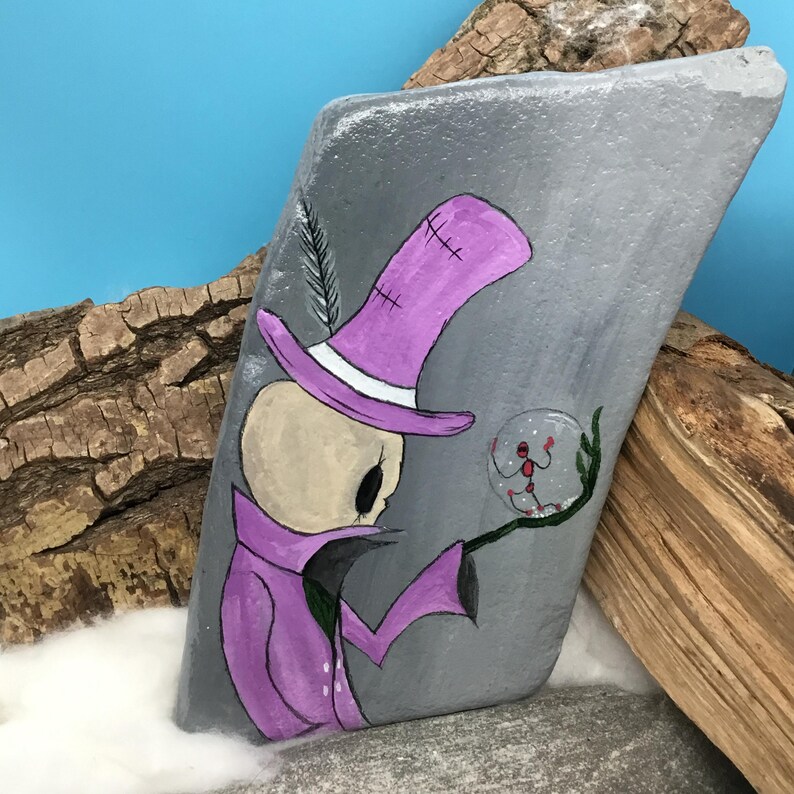 May include: Hand-painted rock art featuring a whimsical skeleton figure in a purple top hat and coat, holding a crystal ball. The artwork is set against a gray background with a blue sky and wood accents, creating a unique decorative piece.
