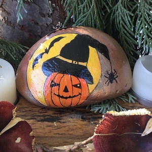May include: A painted rock featuring a yellow moon, a black witch's hat, and a jack-o'-lantern face. The rock is surrounded by greenery and two white candles.