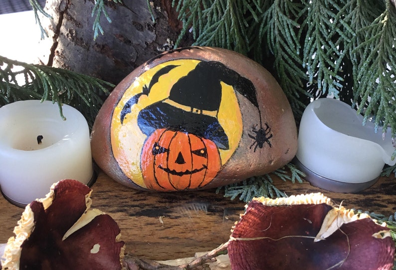 May include: A painted rock featuring a yellow moon, a black witch's hat, and a jack-o'-lantern face. The rock is surrounded by greenery and two white candles.