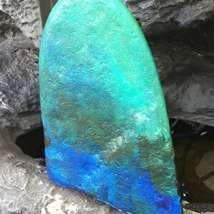 May include: A teal and blue surfboard fin with a textured surface. The fin is painted with a gradient of colours, starting with a dark teal at the top and fading to a bright blue at the bottom.