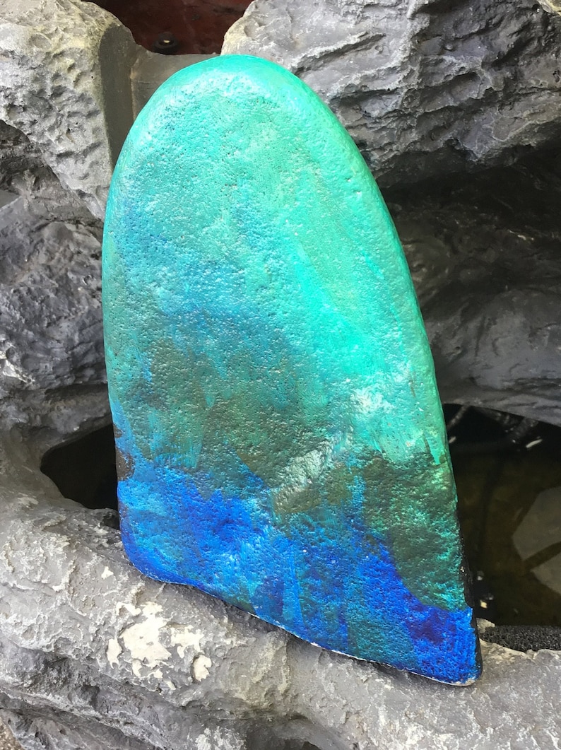 May include: A teal and blue surfboard fin with a textured surface. The fin is painted with a gradient of colours, starting with a dark teal at the top and fading to a bright blue at the bottom.