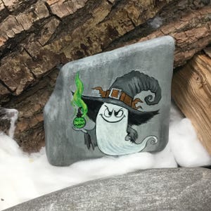 Peut inclure: Une pierre grise peinte &agrave; la main repr&eacute;sente un fant&ocirc;me de dessin anim&eacute; portant un chapeau de sorci&egrave;re avec une ceinture marron. Le fant&ocirc;me tient une bouteille de potion verte avec de la fum&eacute;e verte. L'&oelig;uvre est sur fond de bois et de duvet blanc.