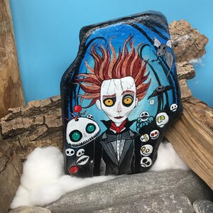Pu&ograve; includere: Arte rupestre dipinta a mano con un personaggio con capelli rossi, occhi gialli e un abito nero. L'opera d'arte include altri personaggi spettrali e uno sfondo blu e nero. La roccia &egrave; esposta con legno e cotone.