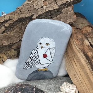 May include: A hand-painted, grey stone features a white owl holding a letter with a red wax seal. The owl is perched on a grey rock, with a blue background and wooden accents. The artwork is detailed and whimsical.