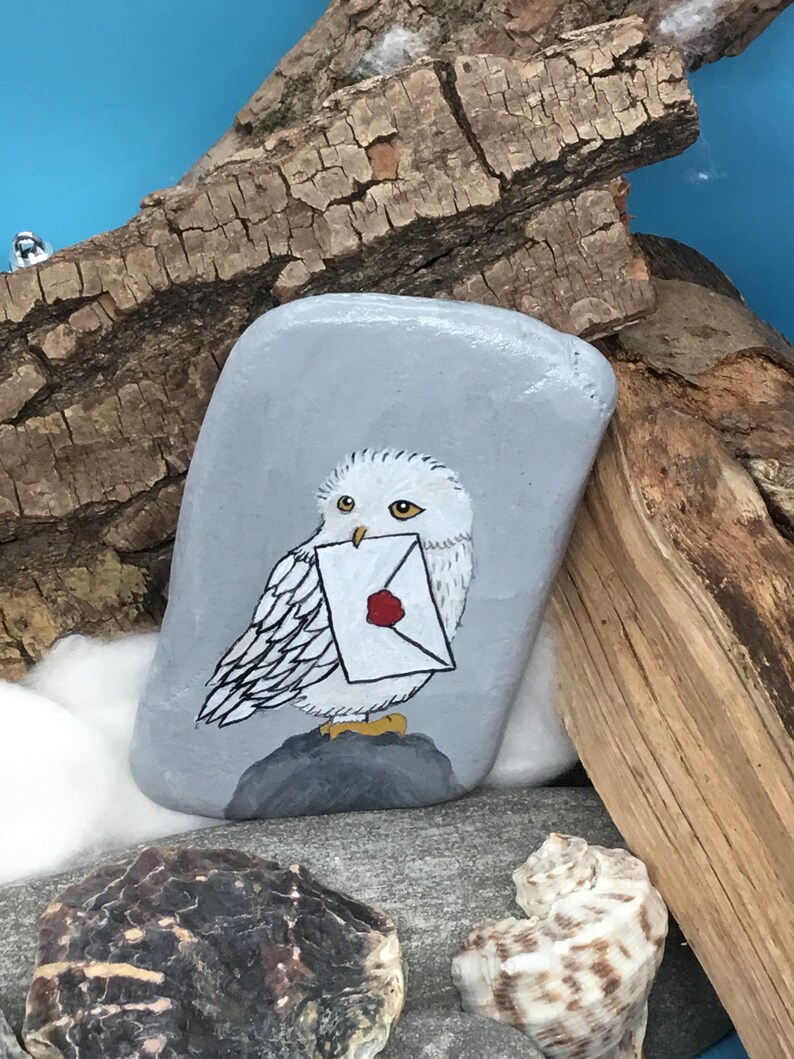 May include: A hand-painted, grey stone features a white owl holding a letter with a red wax seal. The owl is perched on a grey rock, with a blue background and wooden accents. The artwork is detailed and whimsical.