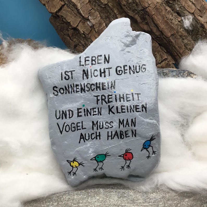 May include: A light gray painted rock featuring black hand-lettered German text that reads: LEBEN IST NICHT GENUG SONNENSCHEIN FREIHEIT UND EINEN KLEINEN VOGEL MUSS MAN AUCH HABEN. Below the text are four small, colorful bird illustrations in yellow, green, red, and blue.