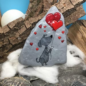 May include: A hand-painted gray rock with a cartoon dog holding two red heart-shaped balloons. The dog is surrounded by smaller red hearts.