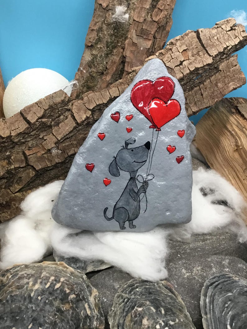 May include: A hand-painted gray rock with a cartoon dog holding two red heart-shaped balloons. The dog is surrounded by smaller red hearts.
