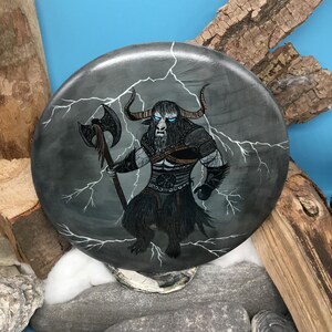 May include: A circular, grey decorative item depicting a mythical beast with bull horns, a long beard, and blue eyes. The creature holds an axe, with white lightning bolts around it. The artwork has a fantasy theme.
