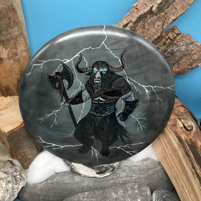 May include: A circular, grey decorative item depicting a mythical beast with bull horns, a long beard, and blue eyes. The creature holds an axe, with white lightning bolts around it. The artwork has a fantasy theme.