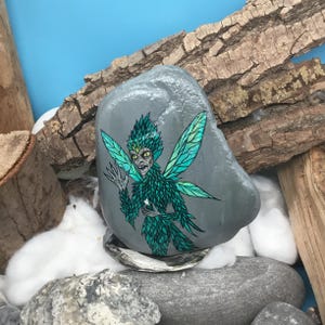 May include: A hand-painted gray stone features a green fairy with detailed wings and a menacing expression. The fairy is adorned with green feathers and holds a small white star. The artwork is set against a backdrop of wood and cotton.