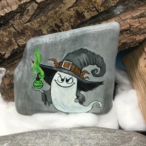 Peut inclure: Une pierre grise peinte &agrave; la main repr&eacute;sente un personnage fant&ocirc;me fantaisiste portant un chapeau de sorci&egrave;re. Le fant&ocirc;me tient une potion verte et a un sourire diabolique. Le chapeau est gris avec une boucle et une garniture marron. L'&oelig;uvre est sur fond de bois et de coton.