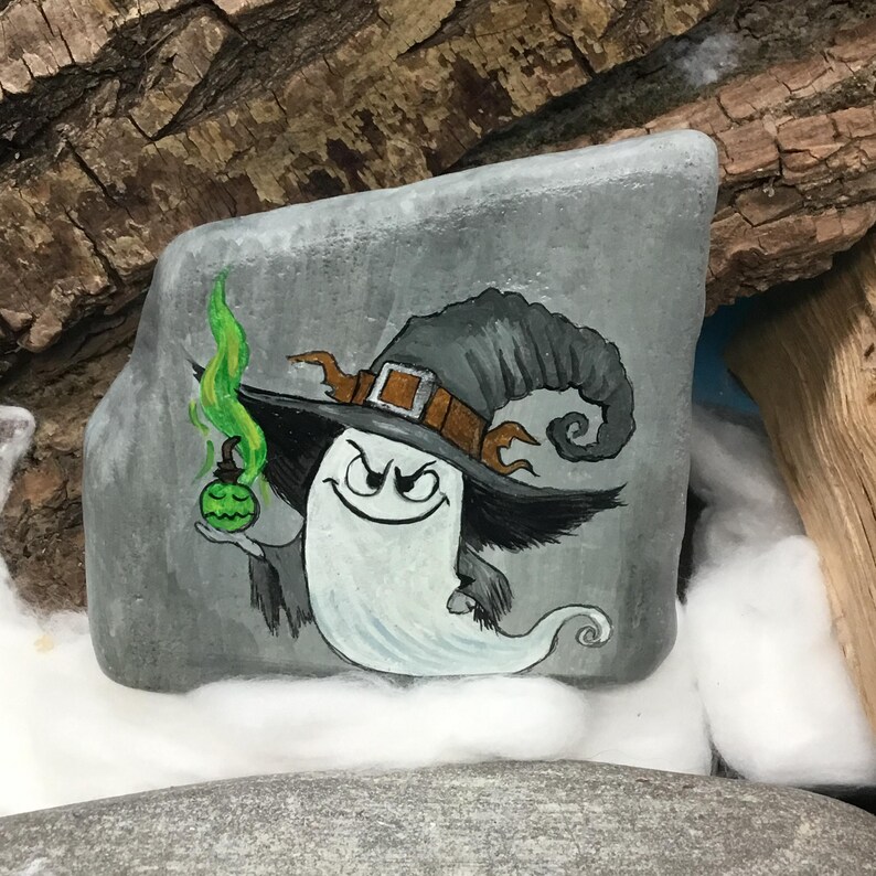 Peut inclure: Une pierre grise peinte &agrave; la main repr&eacute;sente un personnage fant&ocirc;me fantaisiste portant un chapeau de sorci&egrave;re. Le fant&ocirc;me tient une potion verte et a un sourire diabolique. Le chapeau est gris avec une boucle et une garniture marron. L'&oelig;uvre est sur fond de bois et de coton.
