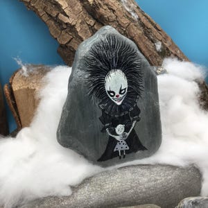 May include: A grey stone painted with a detailed illustration of a clown with wild black hair, a white face, and a red nose, wearing a black ruffled collar and dress, holding a small doll with a white face and a grey dress with black polka dots.