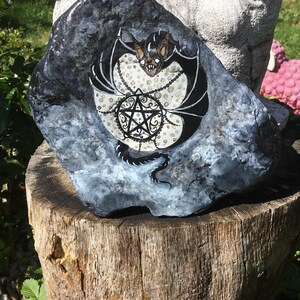 May include: A hand-painted rock featuring a bat design with a pentagram. The bat has black wings and a brown face. The pentagram is black with white accents. The rock is dark gray with white and blue accents. The rock is sitting on a wooden stump.