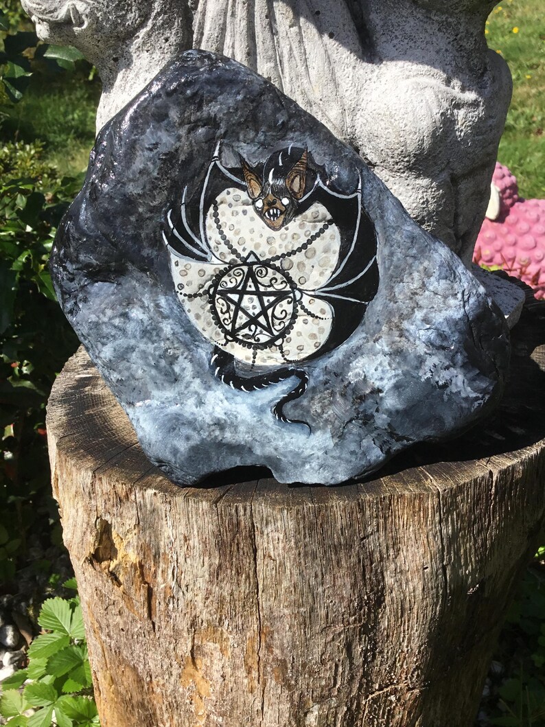 May include: A hand-painted rock featuring a bat design with a pentagram. The bat has black wings and a brown face. The pentagram is black with white accents. The rock is dark gray with white and blue accents. The rock is sitting on a wooden stump.
