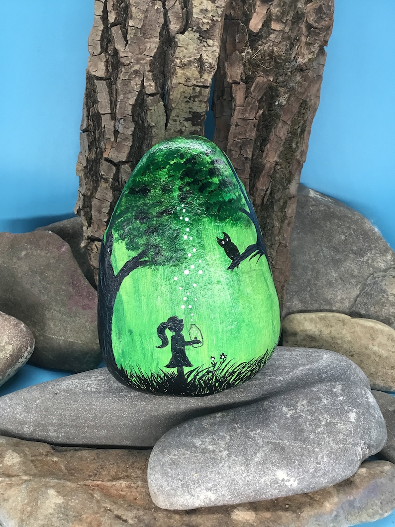 K&ouml;nnte beinhalten: Handbemalter Stein mit der Darstellung eines M&auml;dchens in einem Wald mit einem leuchtenden Glas, einem Baum und einem Vogel. Der Stein ist gr&uuml;n mit wei&szlig;en Akzenten und schwarzen Umrissen.