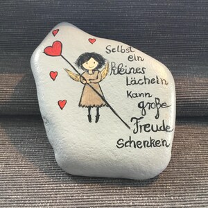 K&ouml;nnte beinhalten: Ein bemalter Stein mit hellblauem Hintergrund zeigt einen Cartoon-Engel, der einen Stab mit Herzspitze h&auml;lt. Rote Herzen sind um den Engel verstreut. Der deutsche Text lautet: "Selbst ein kleines L&auml;cheln kann gro&szlig;e Freude schenken."