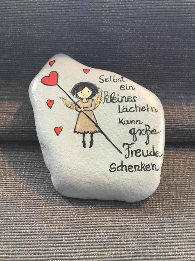 K&ouml;nnte beinhalten: Ein bemalter Stein mit hellblauem Hintergrund zeigt einen Cartoon-Engel, der einen Stab mit Herzspitze h&auml;lt. Rote Herzen sind um den Engel verstreut. Der deutsche Text lautet: "Selbst ein kleines L&auml;cheln kann gro&szlig;e Freude schenken."