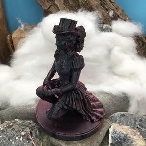 May include: A black and dark red figurine of a person kneeling on a circular base. The figure wears a top hat with goggles, a fitted bodice, and a layered skirt. The surface of the figurine shows a textured, layered finish with red highlights.