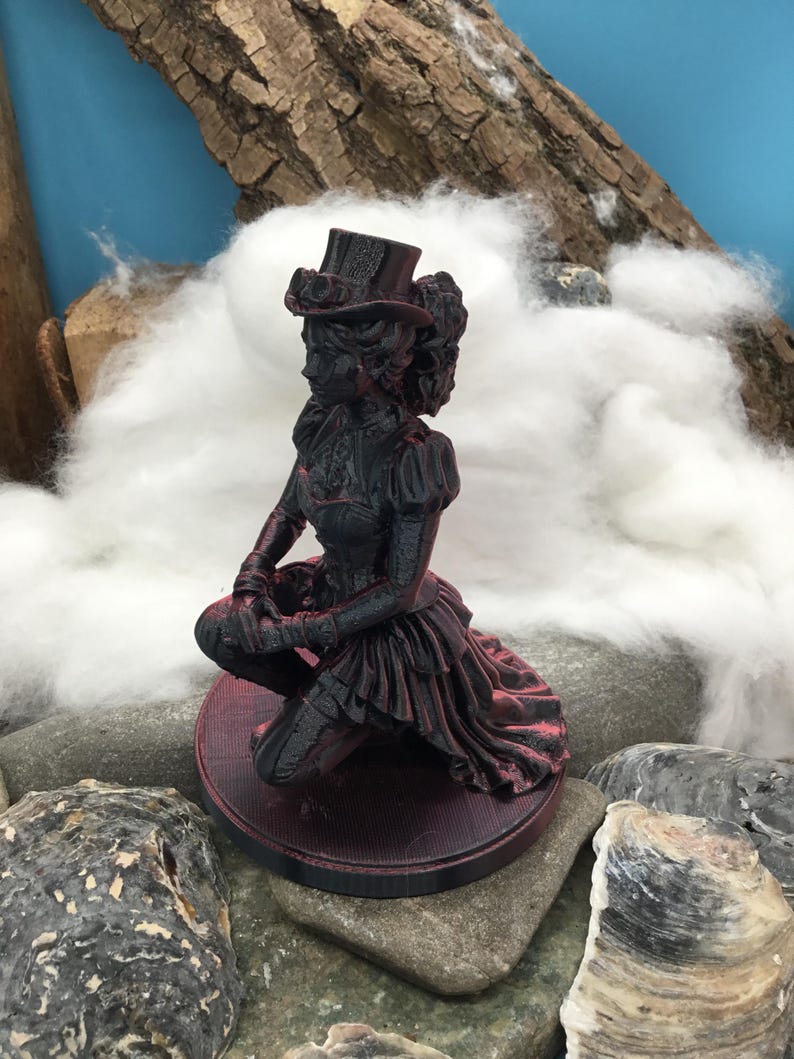 May include: A black and dark red figurine of a person kneeling on a circular base. The figure wears a top hat with goggles, a fitted bodice, and a layered skirt. The surface of the figurine shows a textured, layered finish with red highlights.