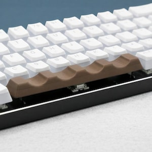 Op de afbeelding: Een compact, wit mechanisch toetsenbord met een zwart frame. Het toetsenbord heeft witte toetsen, een bruine spatiebalk en een gele badeendfiguur aan de linkerkant. Het toetsenbord staat op een wit oppervlak.