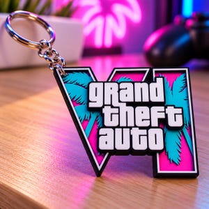 May include: A keychain featuring a black, white, and pink rectangular pendant with the text Grand Theft Auto and a stylized Roman numeral VI. The design includes bright blue palm tree silhouettes against a vibrant pink background with a metal ring.