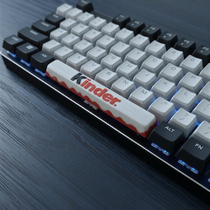 Peut inclure: Un clavier mécanique avec des touches blanches et noires. La barre d'espace porte le mot "Kinder" en rouge et blanc. Le clavier est rétroéclairé en bleu et se détache sur un fond sombre.