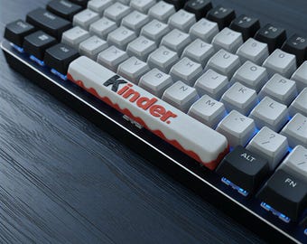 May include: A mechanical keyboard with white and black keycaps. The spacebar features the word "Kinder" in red and white. The keyboard has blue LED backlighting and is set against a dark background.