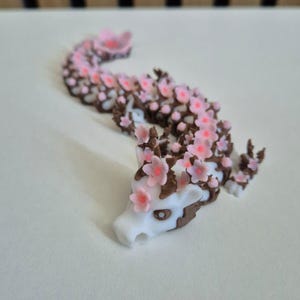 May include: A white and brown articulated dragon figurine featuring a long, segmented body adorned with numerous small, light pink cherry blossom flowers. The dragon has a white head with dark eyes and brown accents along its back and limbs.