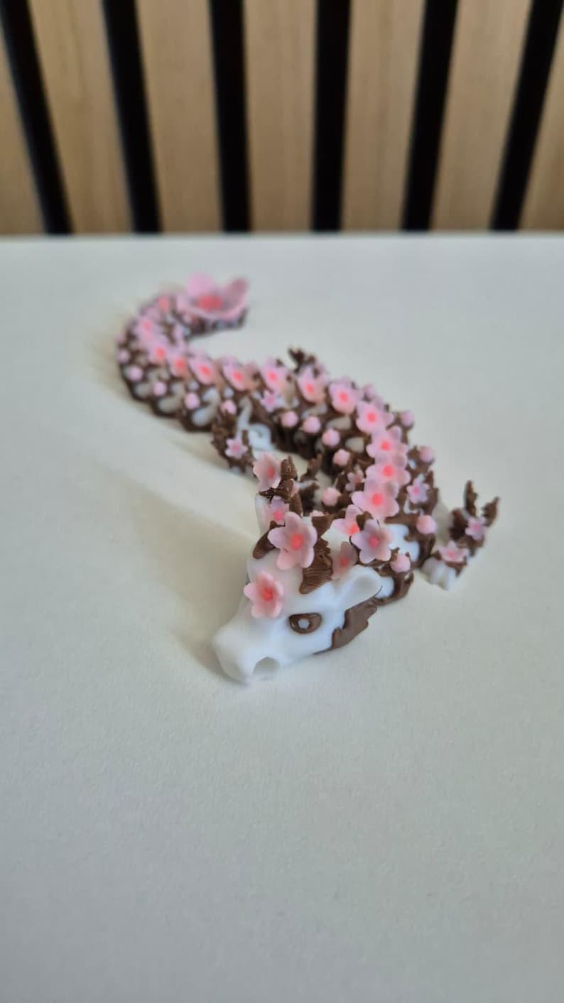 May include: A white and brown articulated dragon figurine featuring a long, segmented body adorned with numerous small, light pink cherry blossom flowers. The dragon has a white head with dark eyes and brown accents along its back and limbs.