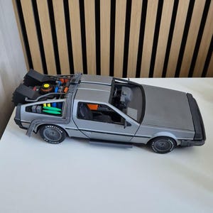 May include: A silver scale model of a sports car featuring a complex rear engine assembly with black, blue, and green components, black wheels with Goodyear branding, and an open cockpit interior with dark seats and a dashboard visible from above.