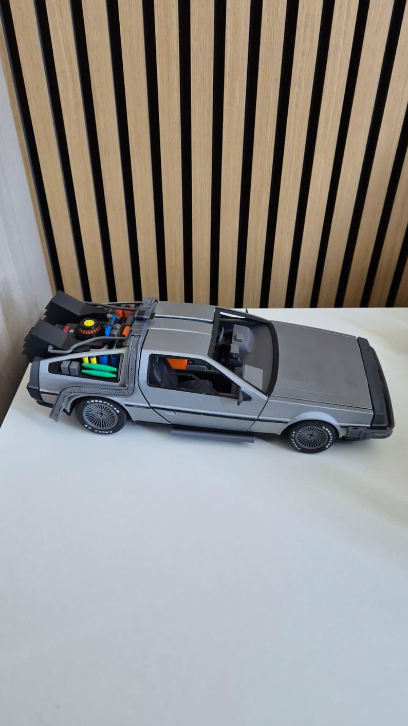 May include: A silver scale model of a sports car featuring a complex rear engine assembly with black, blue, and green components, black wheels with Goodyear branding, and an open cockpit interior with dark seats and a dashboard visible from above.