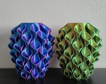 Könnte beinhalten: Zwei 3D-gedruckte Vasen mit einer sich wiederholenden geometrischen, gerüschten, blütenblattartigen Textur. Die linke Vase zeigt einen Farbverlauf von Blau und Violett, während die rechte Vase einen Verlauf von Grün und Gold aufweist.
